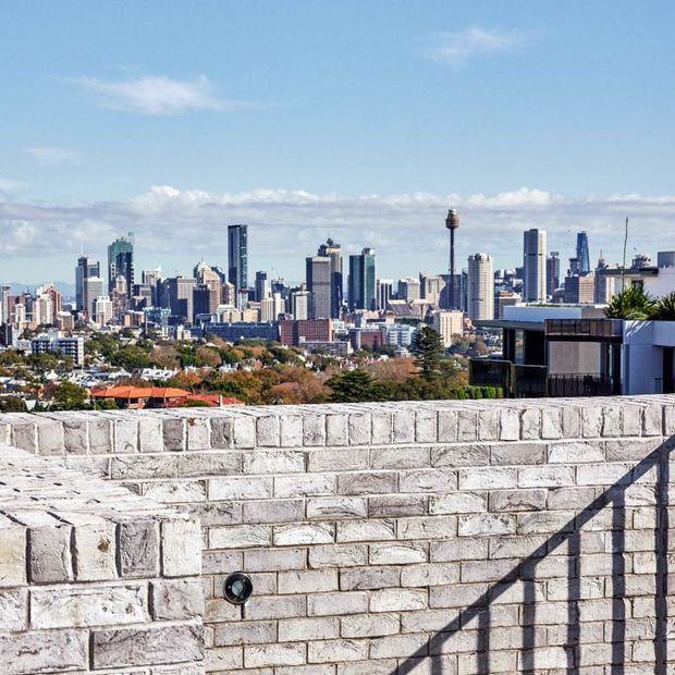 Exterior rooftop or terrace view overlooking a dense urban skyline, with a low light-grey rendered masonry parapet wall dominating the foreground. A modern flat-roof building is visible at right, and the city skyline extends across the background under a bright blue sky.