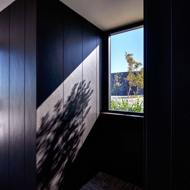 A compact, darkly finished interior corner with floor-to-ceiling vertical timber wall panelling in a near-black finish. A small black-framed window brings in strong sunlight, casting a crisp diagonal beam and tree-shadow pattern across the wall. Dark brick-like flooring is visible at the base, with landscaped greenery and a blue sky seen outside.