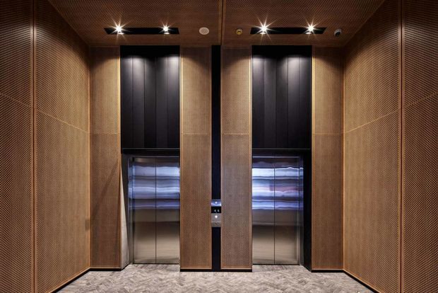 A refined elevator lobby with two elevator doors framed by dark timber-toned perforated wall and ceiling panels. The lifts feature black upper doors and reflective metallic lower panels, while recessed spotlights and linear lighting create a dramatic, warm atmosphere. The floor has a patterned stone or tile finish.