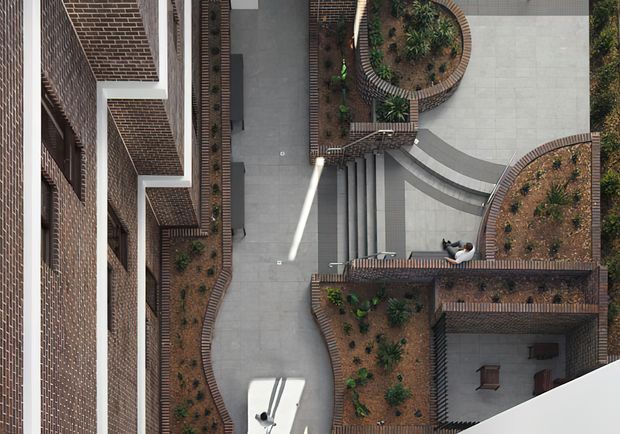 Overhead architectural view of a landscaped urban courtyard framed by red-brick building walls and terraces. Curved brick-edged garden beds, paved walkways, staircases, raised planters, and a small seating area create a sculptural, geometric composition.