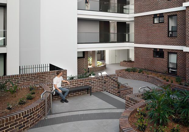 A landscaped communal courtyard and access walkway within a contemporary multi-storey apartment or institutional complex. Curved brick planter walls and integrated brick seating frame broad grey paved ramps and steps with stainless-steel handrails. Red-brown brick facades, white rendered sections, dark-framed windows, balconies, and planted garden beds create a clean, modern urban setting. A person is seated on the built-in bench.