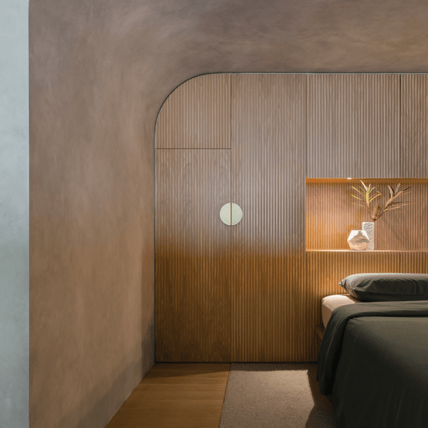 A warm, refined bedroom interior featuring a dark upholstered bed, timber flooring, and a dramatic built-in ribbed timber wall with integrated shelving and concealed storage. The wall treatment curves smoothly into the adjacent plaster wall, while subtle integrated lighting highlights a ceramic vase and dried branch arrangement. The palette is earthy brown, bronze, taupe, and charcoal, creating a calm, intimate atmosphere.
