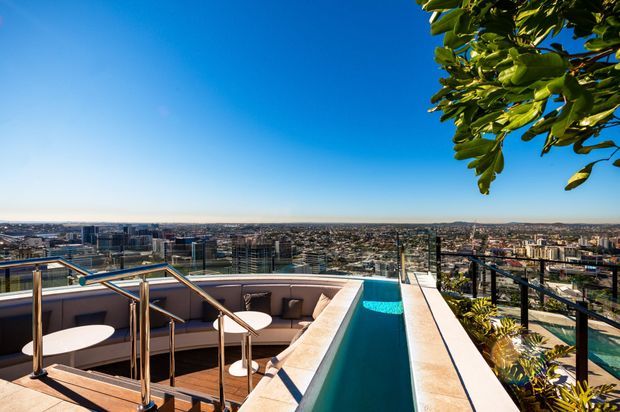 A rooftop terrace overlooking a dense cityscape, featuring a narrow rooftop plunge pool, curved built-in outdoor seating, small round tables, glass balustrades, and metal handrails. Tropical foliage frames the upper right and foreground, with a bright blue sky and expansive urban views beyond.