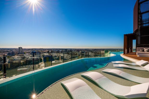 A rooftop or high-rise pool terrace with a long curved infinity-edge swimming pool, submerged sculptural loungers, glass balustrades, and panoramic urban and coastal views. The scene is bright and contemporary, with dark architectural screens and a luxury resort-like outdoor setting.