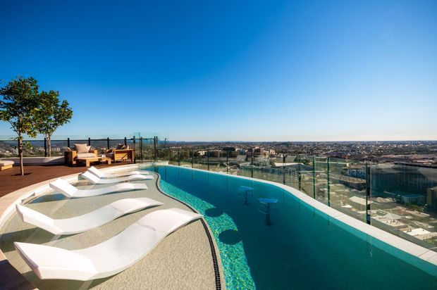 A rooftop or elevated pool terrace with a long turquoise swimming pool, sculptural white sun loungers, glass safety balustrades, and a small outdoor seating area. The view overlooks a dense suburban or urban landscape beneath a clear blue sky.