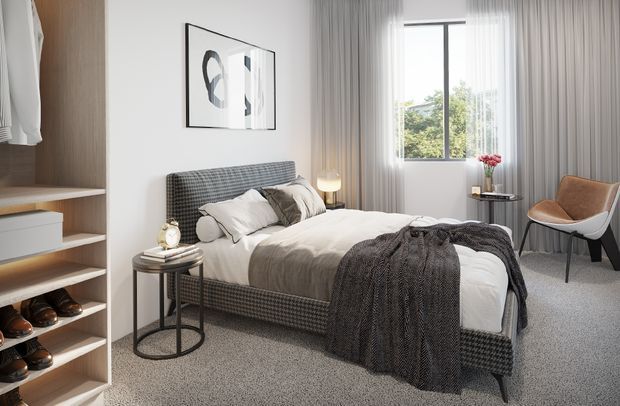 A bright, contemporary bedroom with a neatly made upholstered bed, layered neutral bedding, and a dark textured throw. Built-in open shelving and wardrobe storage line the left wall, while sheer floor-to-ceiling curtains frame a large window. A small round bedside table, bedside lamp, abstract artwork, and a modern accent chair complete the calm, refined interior.