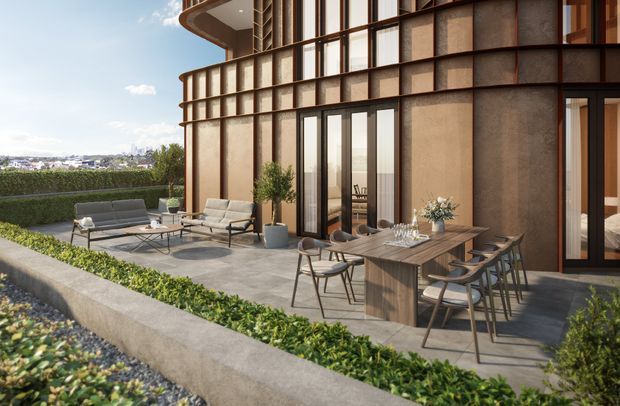 A contemporary outdoor terrace adjoining a curved multi-storey building facade, furnished with a dining table, chairs, lounge seating, and potted plants. The facade combines large glazed openings with dark metal framing and warm brown textured wall panels. Low hedging and a gravelled planted edge define the terrace, with a distant urban landscape visible beyond.