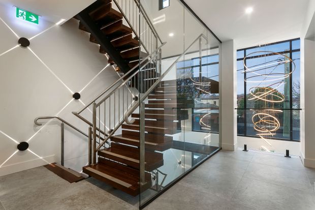 A bright contemporary stairway and landing with dark timber floating stair treads, slim stainless-steel balustrades, and clear glass guarding around a stair void. White walls feature a geometric linear light installation, while tall black-framed glazing overlooks an adjacent building and a sculptural suspended spiral light fitting. The floor is finished in large-format grey tiles or polished concrete.