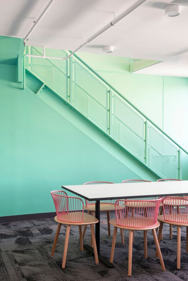 A brightly coloured contemporary interior featuring a mint-green stairwell with perforated metal balustrades, white exposed services and ceiling, and a communal dining or meeting table surrounded by pink wire-frame chairs. Dark patterned carpet contrasts with the pastel green and pink palette.