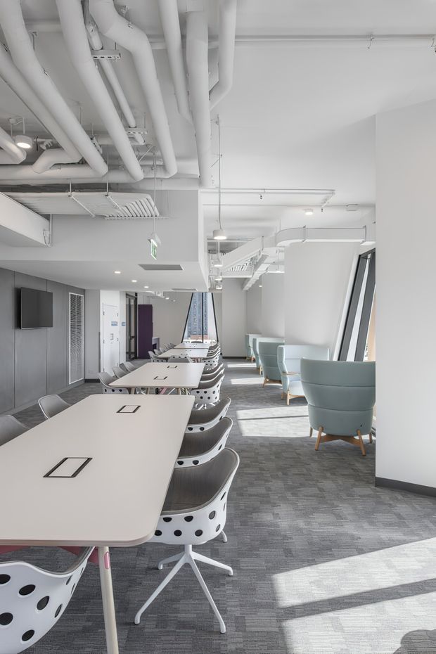 Bright modern commercial office interior with long white collaborative desks, distinctive white chairs with black circular detailing, pale blue upholstered lounge chairs, grey carpet, large angled windows, and exposed white-painted services and ductwork across the ceiling. The open-plan workspace is clean, airy, and contemporary with abundant natural light.