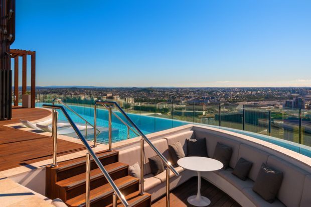 A rooftop or elevated outdoor terrace with a turquoise swimming pool, timber decking and steps, stainless-steel glass balustrades, and a built-in curved outdoor lounge with cushions around a small round table. The terrace overlooks a broad urban landscape under a clear blue sky.