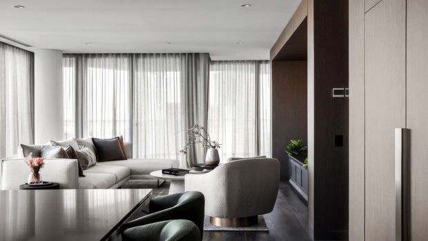 Contemporary open-plan living room with a neutral sectional sofa, rounded lounge chair, dark coffee table, full-height sheer curtains, and a dark timber built-in feature wall with concealed cabinetry. The room is softly lit and refined, with a restrained palette of white, grey, charcoal, and dark wood.