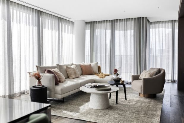 Bright, refined contemporary living room with extensive floor-to-ceiling sheer curtains and glazing, a large neutral sectional sofa, sculptural upholstered armchair, layered round coffee tables, and a textured area rug. The room has a light neutral palette with dark timber flooring and a clean, spacious layout.
