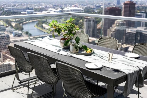 A bright high-rise dining terrace with a long dark dining table set for six, woven outdoor dining chairs, a light linen table runner, tableware, glassware and potted greenery. Full-height glass balustrading frames expansive city and river views in the background.
