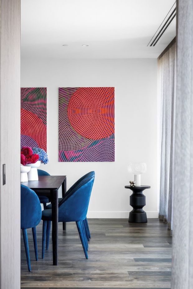 A bright, refined dining area viewed through a doorway, featuring a dark rectangular dining table with vivid deep-blue upholstered chairs, colourful geometric artwork on the white wall, grey-toned timber flooring, full-height sheer curtains, and a small black sculptural side table with decorative objects. Recessed ceiling lights and a linear air-conditioning vent contribute to the clean contemporary fit-out.