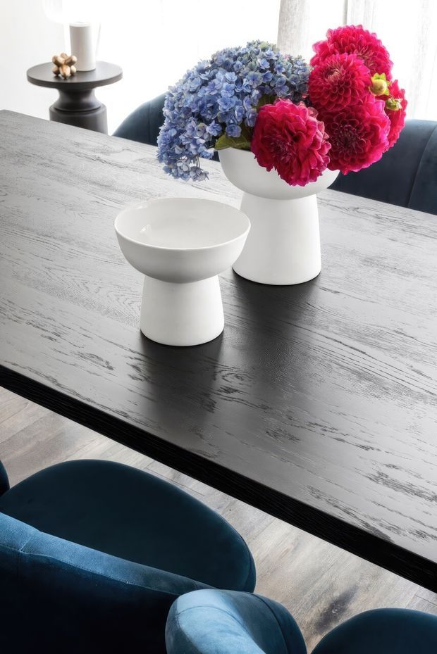 A close-up dining area scene centred on a large dark timber dining table with a textured, distressed-looking surface. Two sculptural white ceramic vessels sit on the table, one holding vivid blue hydrangeas and deep pink flowers. Dark blue upholstered dining chairs, a small black side table, and bright sheer window curtains are visible in the background. The image has a refined, high-contrast contemporary feel with bold floral accents.