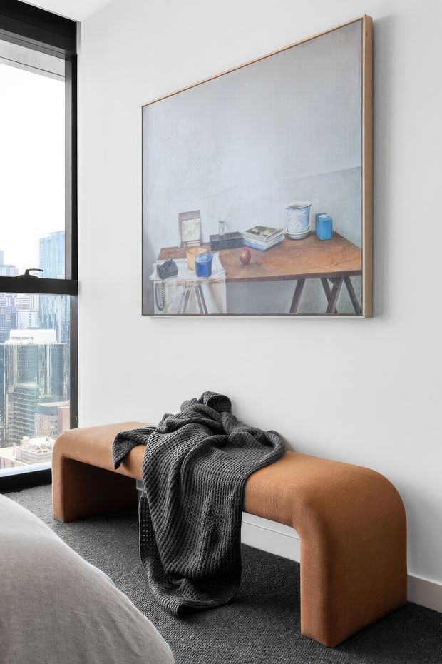A contemporary bedroom corner featuring a sculptural caramel-brown upholstered bench with rounded ends, a dark charcoal knitted throw, and a large framed artwork above. A bed edge is visible in the foreground, while a full-height black-framed window provides city views. The palette is neutral and refined, with dark carpet and white walls.
