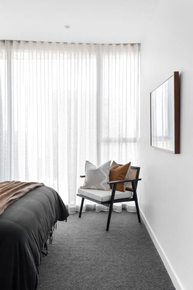 Bright, minimalist bedroom with full-height sheer white curtains covering a large window, a dark upholstered bed with layered neutral throws, and a black-framed lounge chair with white and tan cushions. The room has white walls, grey carpet, a simple framed artwork or mirror, and abundant diffused natural light.