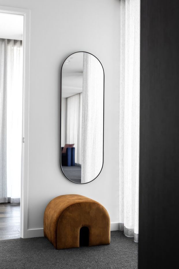 A refined monochromatic interior vignette featuring a tall rounded-rectangle wall mirror with a slim black frame, white walls, sheer floor-to-ceiling curtains, and dark grey carpet. A small sculptural wooden stool or side table sits beneath the mirror, with a dark vertical partition at right and a glimpse into an adjoining room at left. The styling is minimal and contemporary, with strong contrast between pale architectural surfaces, charcoal flooring, and warm timber.