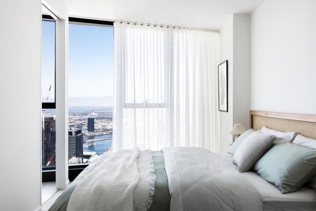 Bright, high-rise bedroom with a large bed dressed in layered white and pale blue bedding. Floor-to-ceiling corner glazing with sheer white curtains provides expansive city and waterfront views. The room has white walls, a light timber and woven-panel bedhead, minimal artwork, and a calm contemporary feel.