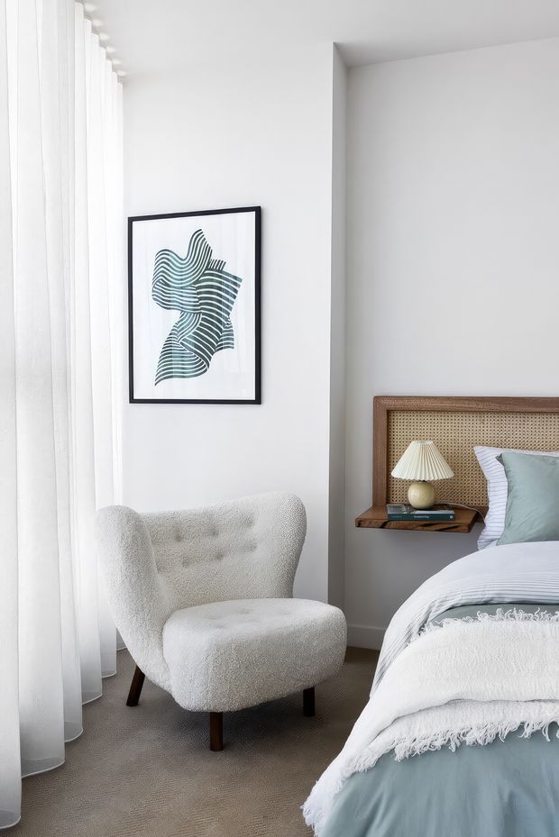 A bright, calm bedroom corner with a partially visible bed, woven cane headboard, pale blue bedding, a textured boucle armchair, sheer full-height curtains, and a small wall-mounted bedside shelf. White walls and soft neutral carpeting create an airy, refined interior, complemented by abstract framed artwork and a small table lamp.