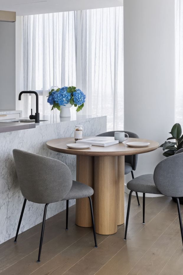 Bright, refined open-plan kitchen and dining nook. A round light-wood dining table with a sculptural pedestal base is surrounded by grey upholstered dining chairs. Behind it, a white marble-look kitchen island with a matte black gooseneck tap, white cabinetry, and a vase of blue hydrangeas sits beneath sheer floor-to-ceiling curtains. The space has pale tiled flooring, white walls, soft natural light, and a clean contemporary feel.