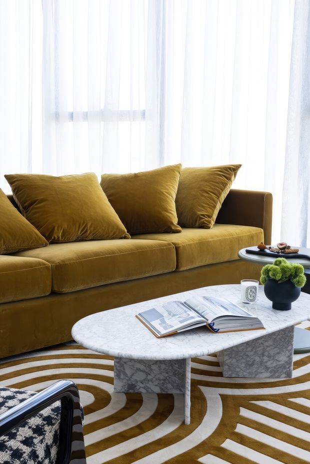 A bright, refined living room vignette featuring a mustard-gold velvet sofa with matching cushions, sheer white curtains, a white marble coffee table with sculptural pedestal legs, and a bold cream-and-mustard geometric rug. A coffee-table book and small decorative objects add a styled, luxurious finishing touch.