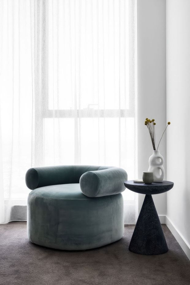 A softly lit minimalist sitting area with a rounded upholstered lounge chair in muted blue-grey, positioned beside a small sculptural black side table. Sheer white curtains cover a large window, while a white sculptural vase and neutral ceramic vessel provide understated decorative accents. The room has white walls, pale trim, and a grey-brown textured carpet.