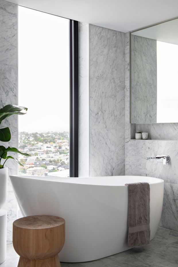 Bright contemporary bathroom featuring a freestanding white bathtub beside a full-height window, grey-white veined marble wall and floor surfaces, a large frameless mirror, and a wall-mounted chrome tap. A wooden stool and grey towel add warmth, while a leafy plant sits by the window overlooking a dense urban neighbourhood.
