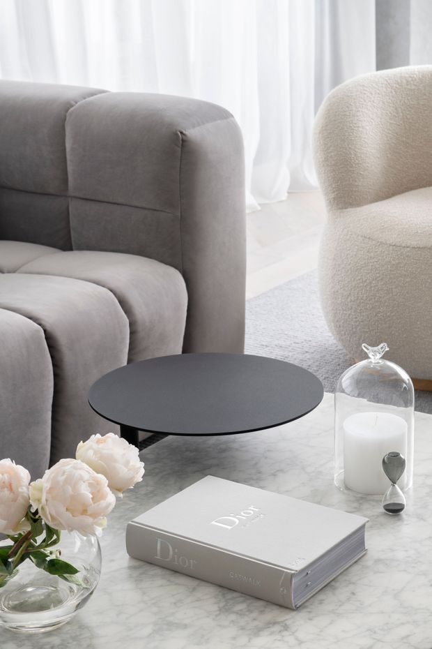 A softly styled contemporary living room vignette featuring a plush grey sofa and rounded cream boucle armchair around a low black circular side table. A white marble coffee table holds a glass candle cloche, hourglass, floral arrangement, and a neutral-toned design book. Sheer white curtains and pale flooring create a bright, refined atmosphere.