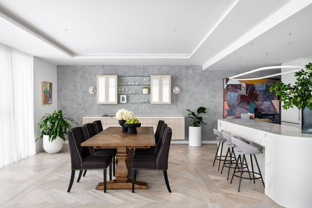 Bright open-plan dining and home bar area with a large timber dining table surrounded by dark upholstered chairs, set against a textured grey feature wall with built-in display cabinetry. A white marble-look bar island with stools occupies the right side, while contemporary artwork, plants, sheer curtains, and layered ceiling details add visual interest. The space has a refined contemporary feel and herringbone-style pale flooring.