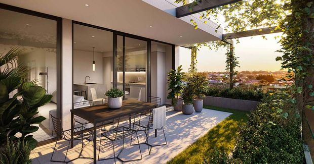 A contemporary outdoor terrace adjoining a glazed interior, with a dark-framed floor-to-ceiling sliding door, dining table and chairs, potted plants, built-in garden edging, and a lush vertical garden. The terrace overlooks a suburban landscape and is shaded by a modern pergola-like overhead frame, creating a warm indoor-outdoor entertaining space.