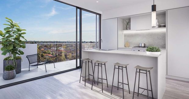 Bright contemporary kitchen and breakfast bar opening onto a balcony through full-height black-framed glazing. The space features a white minimalist kitchen with integrated cabinetry, a pale textured splashback, a slim stone-look benchtop, four simple bar stools, light timber-look flooring, and a potted plant. A broad suburban view is visible beyond the balcony.