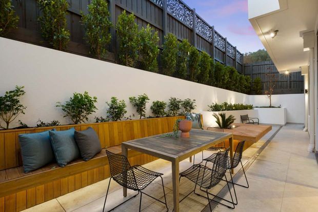 A refined outdoor courtyard dining and lounge area with built-in timber bench seating, a long dining table, wire-frame chairs, tiled paving, raised planting beds, and lush greenery enclosed by rendered walls and decorative metal screening. The space is shown in warm evening light beside a contemporary building facade.