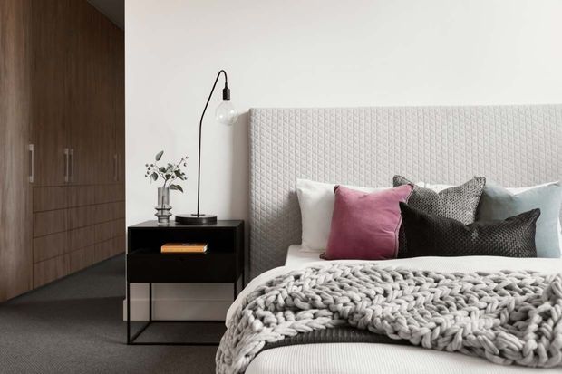 A contemporary bedroom with a large upholstered diamond-quilted headboard, layered pillows, and a chunky knitted throw on the bed. A slim black bedside table and floor lamp sit beside the bed, while dark timber built-in wardrobes line the left side. The room has a restrained palette of white, grey, black, and muted pink with soft, refined styling.