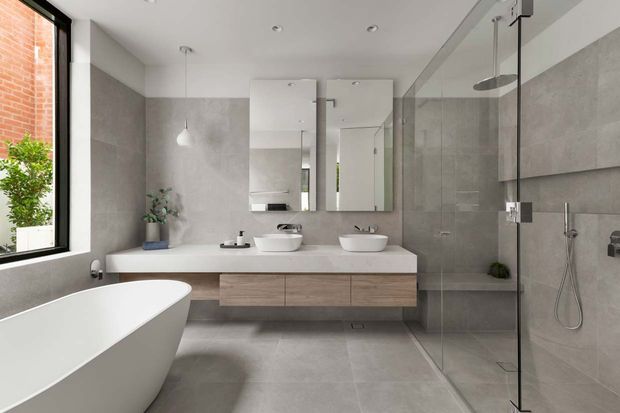 A contemporary bathroom with a freestanding white bathtub, double floating vanity, twin vessel basins, large mirrors, and a frameless glass walk-in shower. The room features large-format grey wall and floor tiles, pale timber vanity cabinetry, recessed ceiling lights, and a full-height window beside exposed brick and greenery.