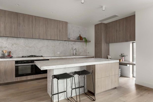 Contemporary open-plan kitchen with full-height wood-look cabinetry, a large pale stone island with two black metal bar stools, matching stone splashback and benchtops, integrated oven and cooktop, timber flooring, and a small adjoining workspace visible to the right. The space is bright, clean, and neutrally toned.