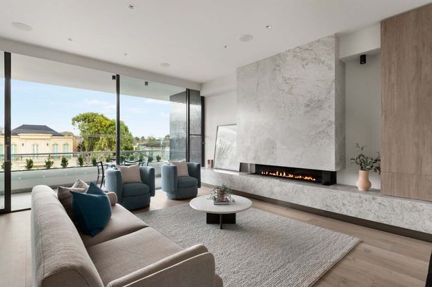 Contemporary living room with floor-to-ceiling glazing, a large pale stone fireplace feature wall, timber-toned built-in cabinetry, a neutral sectional sofa, blue lounge chairs, and a low round coffee table. The room opens to a balcony and overlooks neighbouring homes and greenery.