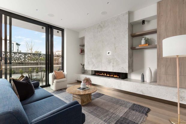 Bright contemporary living room with a blue sectional sofa, textured neutral rug, timber flooring, and a long linear fireplace set into a large pale stone feature wall. Built-in timber shelving and cabinetry flank the fireplace, while full-height black-framed glazing opens to a balcony or terrace.