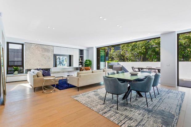 Bright, spacious open-plan living and dining area with floor-to-ceiling glazing, pale timber flooring, a large patterned rug, contemporary upholstered sofas and dining chairs, and sleek built-in cabinetry. The room opens visually to a planted outdoor courtyard or garden.