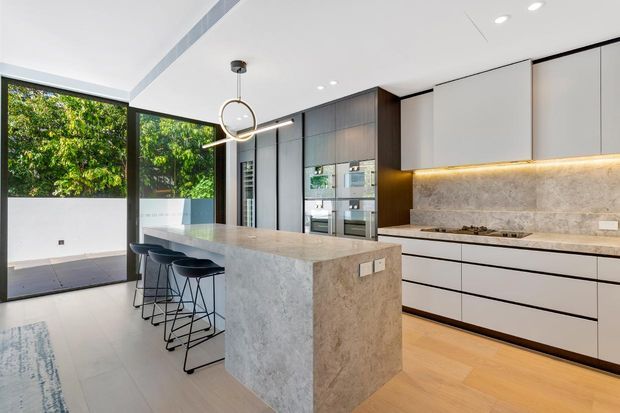Contemporary open-plan kitchen with a large stone island and three black bar stools, dark integrated cabinetry, pale upper cabinets, and a matching stone splashback. Floor-to-ceiling glazing opens to a leafy outdoor area, while recessed lighting and a circular pendant provide a clean, refined finish.