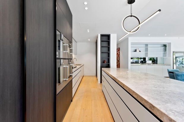 Contemporary open-plan kitchen viewed along a galley-style run toward a large central island. The space features tall dark-wood cabinetry with integrated appliances, pale flat-panel island cabinetry, a light stone benchtop, warm timber flooring, white walls and ceiling, open display shelving, and a sculptural linear pendant light. A living area is visible beyond the kitchen.
