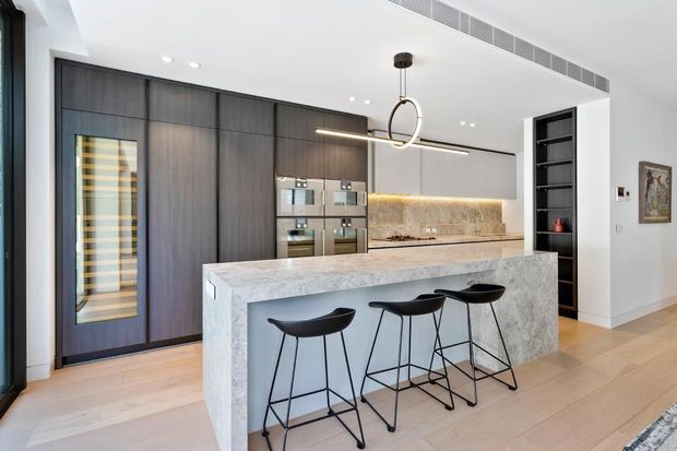 Contemporary open-plan kitchen featuring a large stone island with three black bar stools, dark timber floor-to-ceiling cabinetry, integrated stainless-steel appliances, a warm illuminated stone splashback, pale timber flooring, and a linear ring pendant light. A recessed open shelving unit and adjacent hallway are visible to the right.