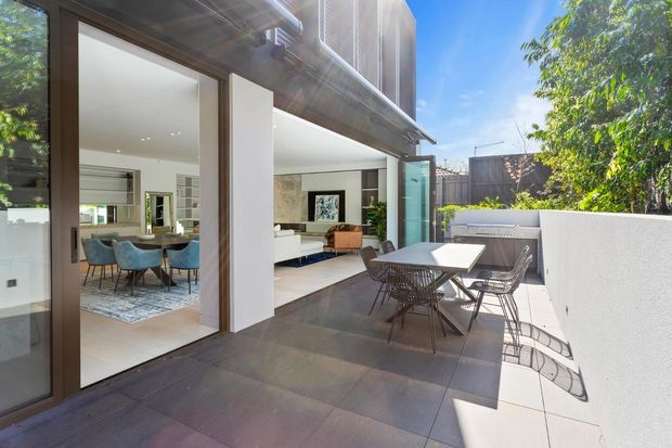 Bright contemporary indoor-outdoor living space viewed from a tiled terrace. Large glazed sliding doors open onto a combined living and dining area with white walls, dark timber-framed glazing, contemporary furniture, and a partly visible kitchen. The terrace includes an outdoor dining table and chairs, while a planted garden and boundary fencing are visible beyond.