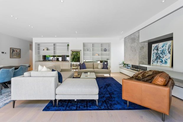 Bright contemporary open-plan living room with a large white sectional sofa, blue patterned rug, tan leather armchair, built-in shelving, and a wall-mounted fireplace with a textured stone feature wall. A dining area is visible to the left, with abundant recessed lighting and neutral finishes accented by deep blue furnishings.