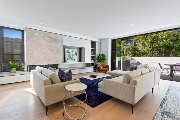 Bright contemporary living room with two cream upholstered sofas arranged around a low coffee table and deep blue rug. The room features pale timber flooring, a white built-in media wall with a large stone-look feature panel, recessed lighting, and extensive glazing opening onto a paved outdoor terrace. Neutral furnishings are accented by navy textiles and indoor plants.