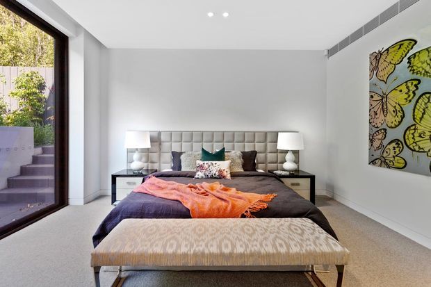 Contemporary bedroom with a centered upholstered bed, tufted headboard, matching bedside tables and lamps, a patterned bench at the foot of the bed, pale carpet, and white walls. Large dark-framed glazing opens to an exterior stairwell, while a colorful butterfly artwork provides a feature accent.