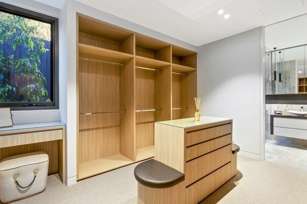 A bright, contemporary walk-in wardrobe with extensive built-in timber-look cabinetry, open hanging bays, shelving and a central drawer island with a cushioned stool. A window seat and integrated storage sit beneath the window, while a glazed opening reveals an adjacent bathroom with a vanity. The room has light carpet, white walls and ceiling, recessed lighting, and dark-framed glazing.