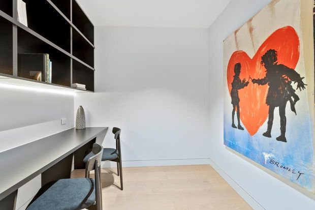 A compact modern home office or study with a built-in grey desk, shelving, and seating along one wall. Black open shelving provides strong contrast against white walls, while pale timber flooring adds warmth. A large colourful artwork with silhouetted figures and a red heart forms the main feature on the opposite wall.