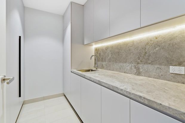 A compact, modern galley-style kitchen or kitchenette with full-height light-grey cabinetry, a pale stone benchtop and matching stone splashback. Integrated warm under-cabinet lighting highlights the textured stone surface, while a stainless-steel sink and minimalist tap sit at the rear. The space has pale tiled flooring, white walls and a clean, streamlined appearance.